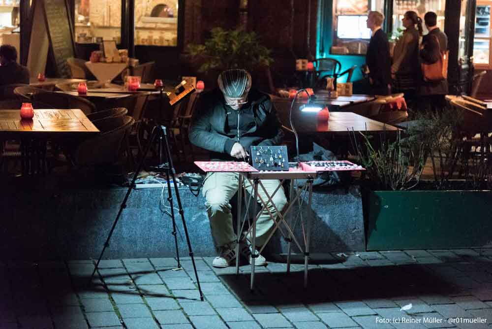 Ein Mann sitzt an einem Tisch und ist ganz in seine Tätigkeit der Schmuckherstellung vertieft. Er sitzt auf einem Platz. Ringsum ist es dunkel. Sein Platz wird von einer Lampe erleuchtet. Im Hintergrund laufen ein paar Menschen im Schein von Laternen.