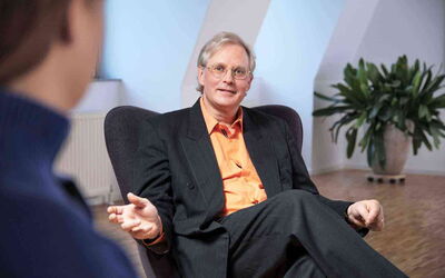 Reiner Müller ist Angsttherapeut und Coach und spezialisiert auf die Unterstützung bei Präsentationsangst. Auf diesem Bild ist er sitzend zu sehen, wie er etwas erklärt.