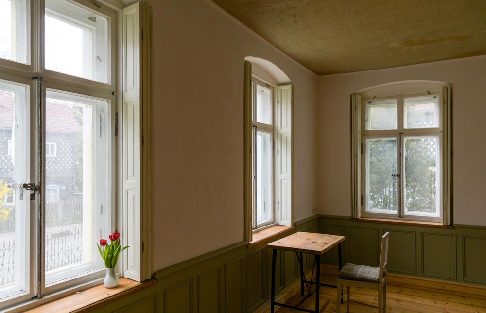 Ein heller, minimalistischer Raum mit großen, minimalistischen Fenstern. Auf der Fensterbank steht eine Vase mit roten Tulpen. In der Ecke des Raumes befindet sich ein Holztisch mit einem schlichten Stuhl, ideal für ruhige Momente oder kreatives Arbeiten.