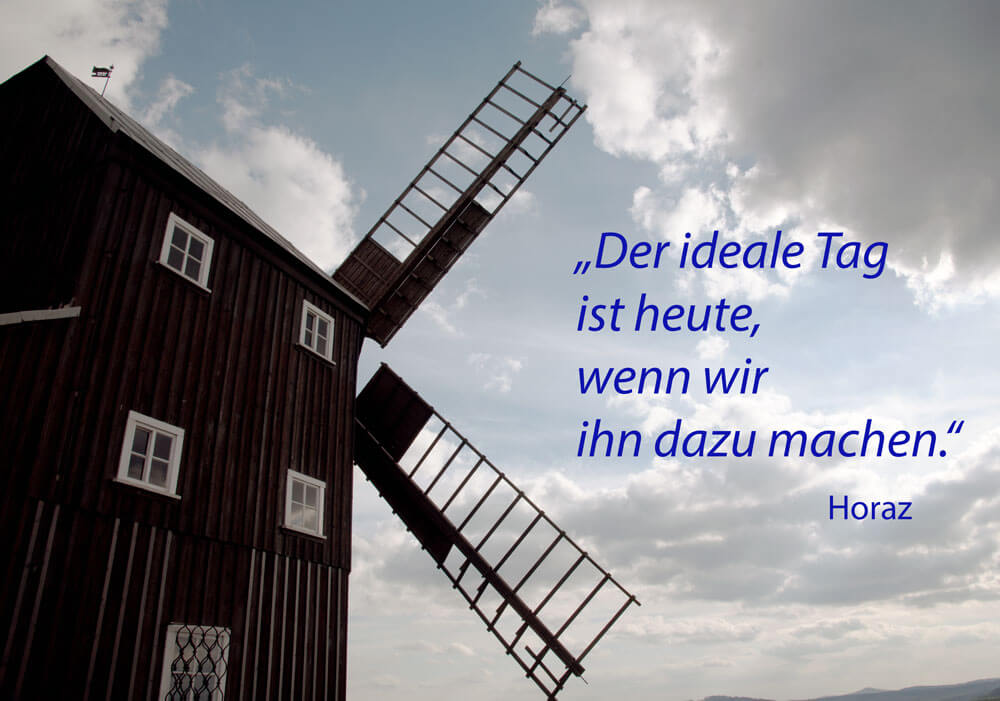 Auf dem Foto ist eine Windmühle vor einem wolkenbedeckten Himmel zu sehen. Auf dem Bild steht der Spruch: "Der ideale Tag ist heute, wenn wir ihn dazu machen." Horaz