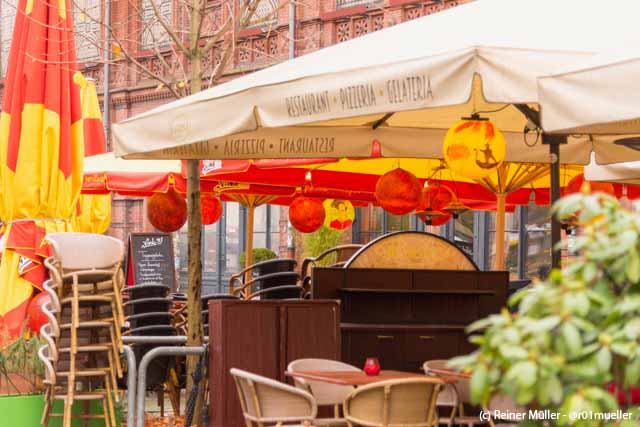 Ein Straßenrestaurant mit bunten Sonnenschirmen und Lampions. Auf einem ist eine Frau, auf einem anderen ein Gondoliere abgebildet.