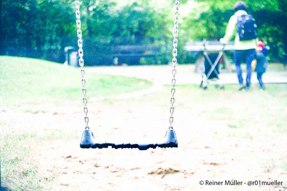 Eine leere Schaukel auf einem Spielplatz. Ein kleines Kind läuft an der Hand ihrer Mutter weg. Es schaut zurück zur Schaukel.